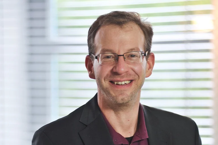 Ein Mann mit Brille lächelt in die Kamera - Im Hintergrund sieht man verschwommen eine Jalousie